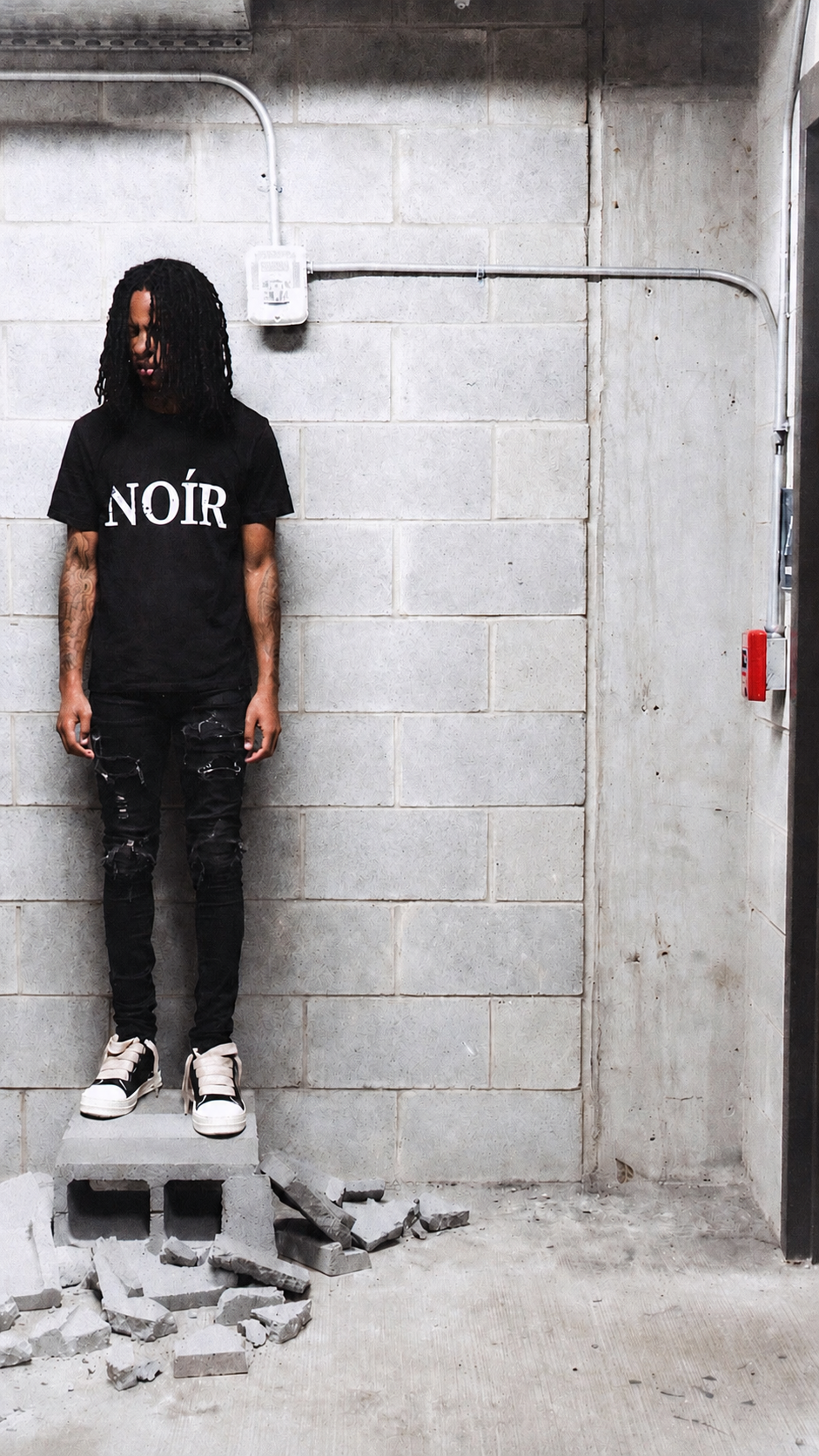 Vendetta Noír luxury streetwear campaign in Chicago. A person stands on a concrete block against a gray cinderblock wall, wearing a black graphic T-shirt with the word 'NOIR' in white, black pants, and white sneakers.